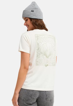 Kobieta w szarej czapce, kremowym T-shircie z grafiką muszli i kwiatów z tyłu oraz napisem "Roxy The Heart of Surfin", ubrana w szare dżinsy.