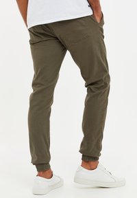 Pantalons tapered vert olive avec des poignets élastiques et des poches arrière, associés à des baskets blanches. Tissu en mélange de coton, texture lisse.