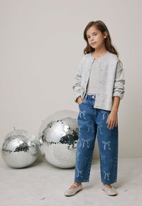 Grå stickad cardigan, ljusgrå topp, jeans med vita rosettmönster och glittriga ballerinaskor, mot bakgrund av reflekterande discokulor.