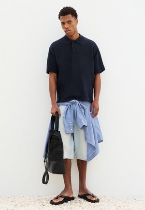 Homme portant un polo bleu marine et un short en denim clair avec une chemise rayée nouée autour de la taille, tenant un sac tressé noir et portant des sandales noires.