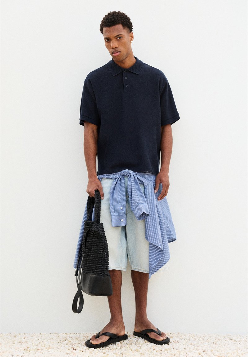 Homme portant un polo bleu marine et un short en denim clair avec une chemise rayée nouée autour de la taille, tenant un sac tressé noir et portant des sandales noires.