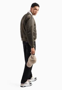 Giubbotto bomber oliva con dettagli ricamati, colletto bianco, jeans neri, cappello beige in mano e scarpe da ginnastica multicolori. Texture liscia.