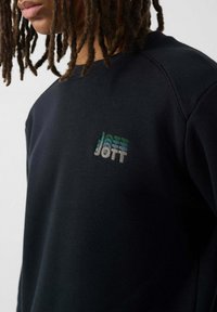 Sudadera azul marino de tejido suave con un logo "JOTT" multicolor en el pecho. El diseño incluye cuello redondo y mangas raglán.