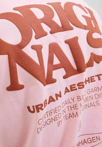 Roosa puuvillane T-särk, millel on suur pruun kiri. Tekst sisaldab sõnu "ORIGINALS" ja "URBAN AESTHETIC," sujuva ja tekstuurita viimistlusega.
