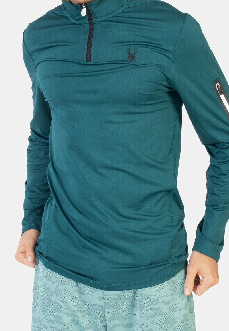 Camiseta deportiva de manga larga en color verde azulado con cuello alto, cierre con cremallera y logo de araña. Presenta una textura suave y un diseño ajustado.