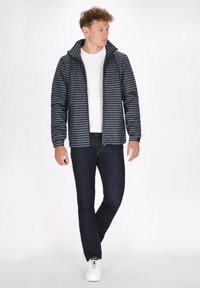 Gestreepte pufferjack in marineblauw met horizontale lichtblauwe lijnen, voorzien van een hoge kraag en ritssluiting, gecombineerd met donkere spijkerbroeken en witte sneakers.