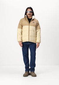 Beige en crèmekleurige gewatteerde jas met een rits aan de voorkant, voorzien van een kraag gevoerd met nepbont, gedragen met blauwe jeans en bruine schoenen.