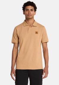 Polo shirt color panna con una texture morbida, maniche corte, colletto classico e un piccolo patch con logo marrone sul petto sinistro. Indossato con pantaloni corti neri.