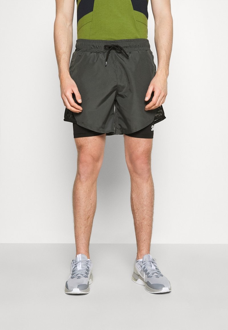 Homme portant un short de course noir superposé, un t-shirt vert et noir, et des chaussures de sport grises, debout devant un fond blanc uni.