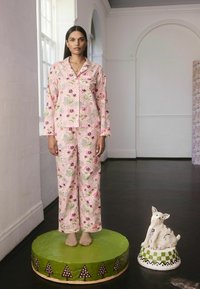 Květinový pyžamový set v světle růžové barvě s vícolorovými květinovými vzory, dlouhými rukávy a volnými kalhotami. Modelka stojí na kulaté zelené platformě.