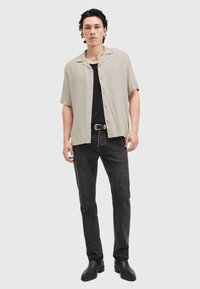 Camisa de botones de manga corta color beige claro, camiseta sin mangas negra, jeans negros y botas de tobillo negras, de pie contra un fondo liso.