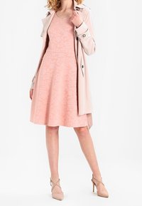 Kvinna som bär en knälång rosa blommig klänning, en beige trenchcoat draperad över axlarna och nude högklackade skor med ankelsnören.