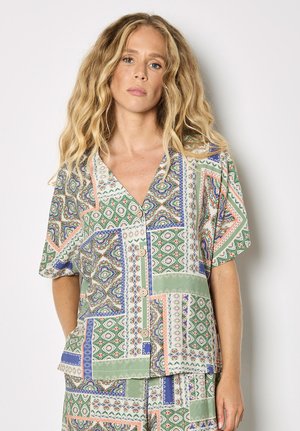 Kobieta o długich kręconych blond włosach, ubrana w krótką koszulę na guziki i pasujące krótkie spodenki z zielonym, niebieskim i beżowym wzorzystym materiałem.