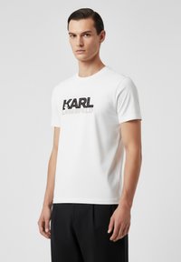 Camiseta blanca de manga corta hecha de tela suave, con un logo gráfico en negro y plata "KARL LAGERFELD" en la parte delantera.