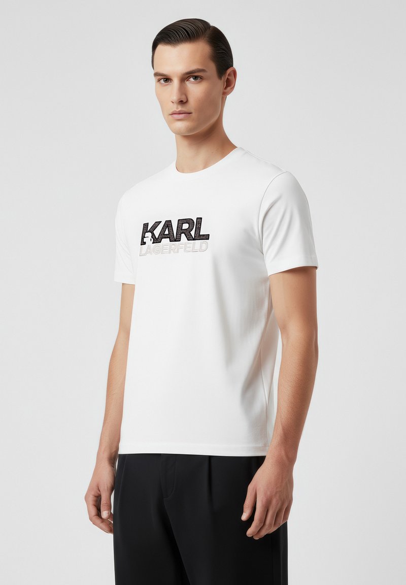 Camiseta blanca de manga corta hecha de tela suave, con un logo gráfico en negro y plata "KARL LAGERFELD" en la parte delantera.