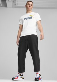 Vit t-shirt med blå och gul Puma-logotyp, kombinerad med svarta baggy jeans och färgglada sportiga sneakers med röda, blå och gula accenter.