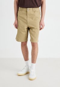 Beige cotton shorts with a straight cut, featuring a front button closure, side pockets, and rolled cuffs, paired with white sneakers and socks.