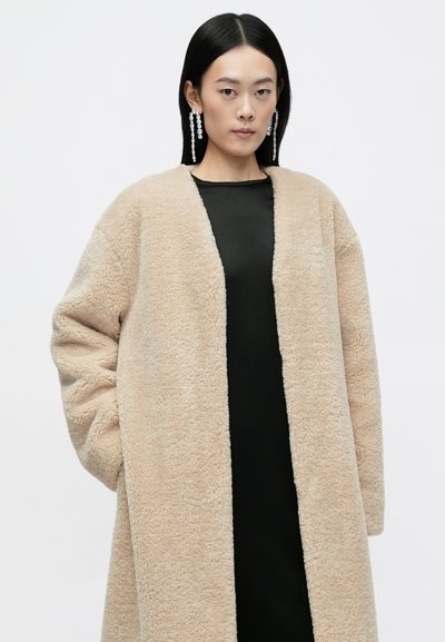 Beige műszőrme kabát, nyitott elülső résszel, hosszú ujjal és puha textúrával, egy fekete ruhára véve. A kabáton nincsenek látható záródások.