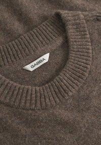 Brauner Strickpullover mit einem gerippten Kragen, der eine weiche Textur aufweist. Enthält ein Etikett mit "GABBA" in fettem schwarzen Text.