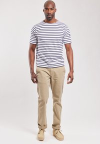 Camiseta de hombre de manga corta, a rayas blancas, con cuello redondo; combinada con chinos beige claro y zapatos marrones, de pie frente a un fondo neutro.