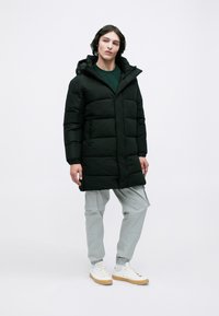 Abrigo de plumas negro con capucha, que presenta secciones acolchadas y bolsillos con cremallera. Llevado sobre una camiseta verde y pantalones cargo grises.