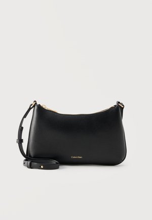 Sac à bandoulière en cuir noir Calvin Klein avec fermeture éclair dorée et sangle réglable sur fond clair uni.
