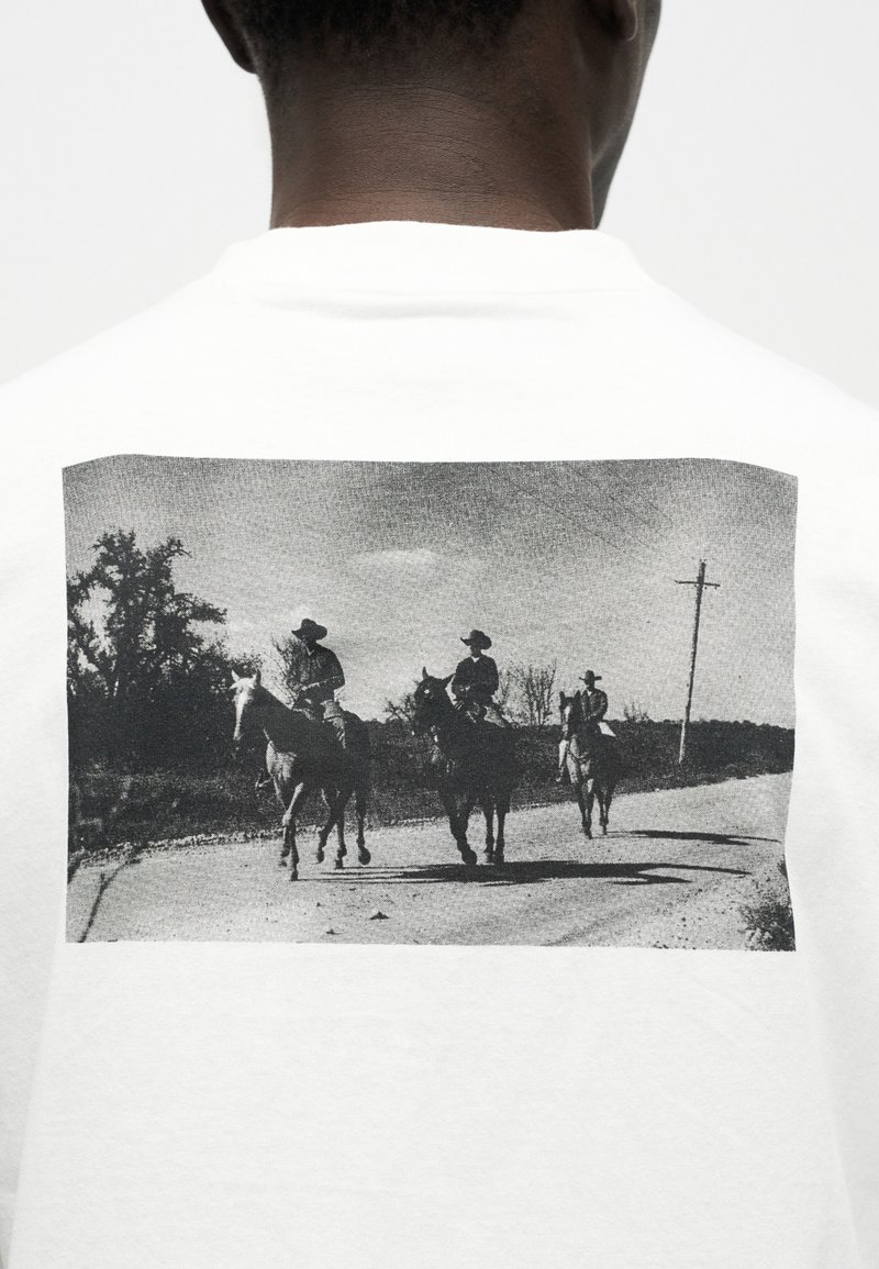 Trois cow-boys chevauchant des chevaux sur une route rurale avec des arbres et un poteau électrique en arrière-plan, illustrés au verso d'un T-shirt blanc.