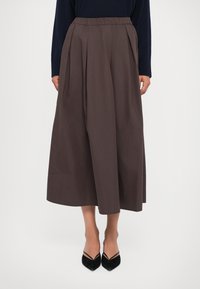 Vrouw in een hoge taille, wijde bruine rok, gecombineerd met zwarte puntige hakken en een marineblauw langsliepend shirt, staande tegen een witte achtergrond.