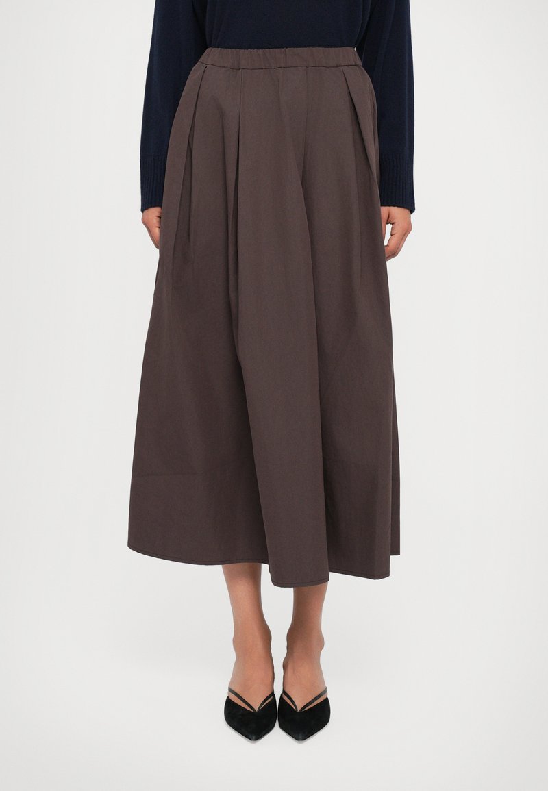 Vrouw in een hoge taille, wijde bruine rok, gecombineerd met zwarte puntige hakken en een marineblauw langsliepend shirt, staande tegen een witte achtergrond.