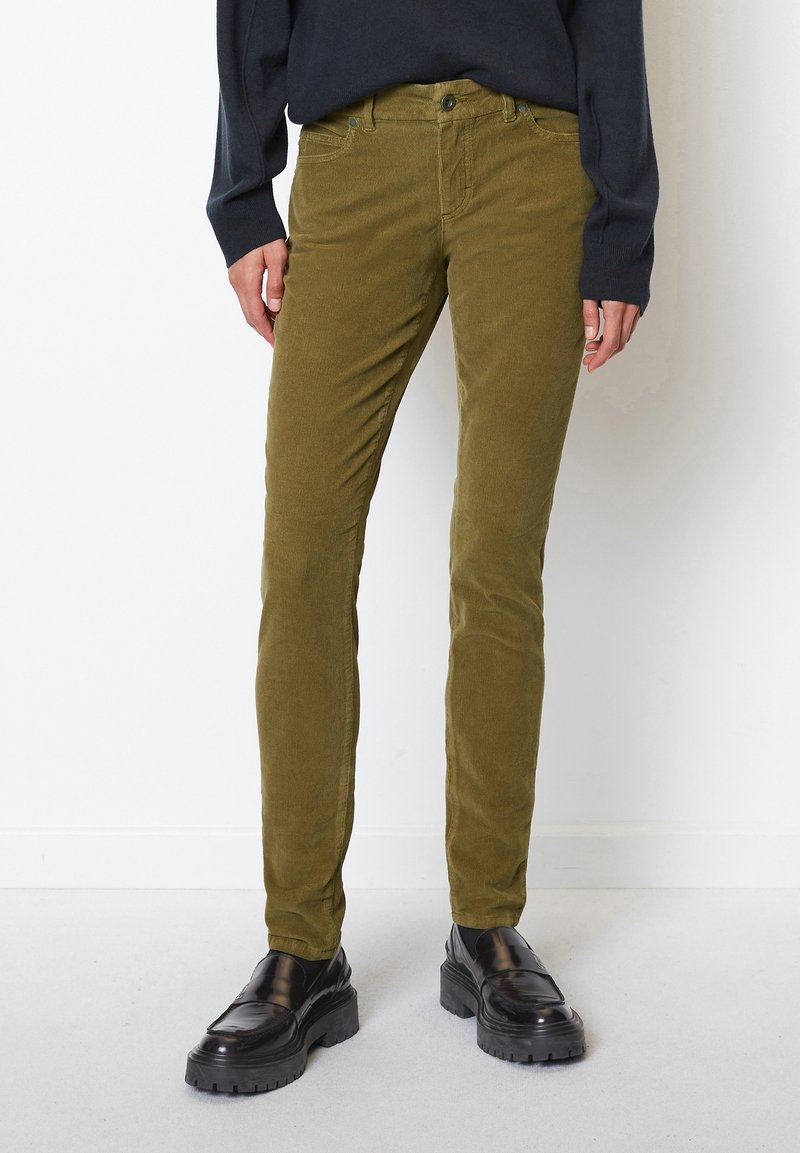 Personne portant un pantalon en velours côtelé vert olive moulant, des mocassins noirs épais et un haut à manches longues foncé, debout contre un mur blanc.