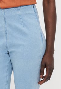 Lyseblå denimbukser med høy liv og sidelommer, samt subtile sømdetaljer, kombinert med en oransje ribbestrikket topp.