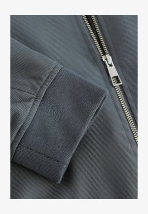 Gray, textured jacket fabric with ribbed cuffs, metallic zipper, and detailed stitching. Smooth surface with a structured design.