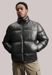 Melna pūkainā jaka ar augstu apkakli, ar quiltēta dizaina, spīdīgu tekstūru un priekšējo rāvējslēdzēju. Modelis stāv ar rokām kabatās.