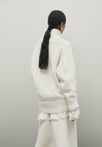Pull blanc confortable avec un col haut et un ourlet côtelé, fabriqué en matériau doux. Porté par-dessus un haut en couches de couleur claire.