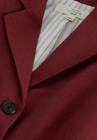 Blazer en lin rouge foncé avec bouton noir, revers cranté et doublure intérieure rayée blanche affichant l’étiquette de la marque.