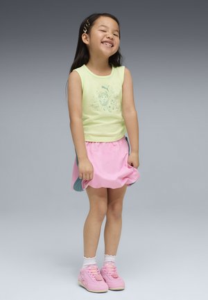 Niña sonriente con camiseta sin mangas amarilla, falda rosa con los lados azules, calcetines blancos, zapatos rosas y una pinza para el pelo, de pie sobre un fondo gris.