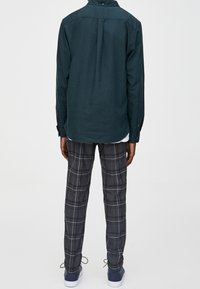 Donkergroene langemouwen shirt met een geknoopte kraag, gecombineerd met grijs geruite broek en blauwe sneakers. Duidelijke katoenachtige textuur zichtbaar.