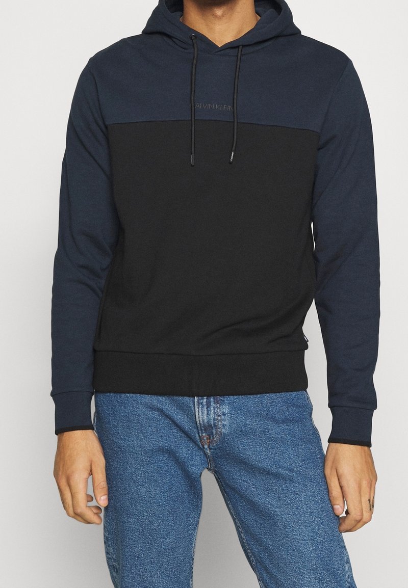 Homme portant un sweat à capuche bicolore bleu marine et noir avec cordons et un jean bleu, debout les bras détendus le long du corps.