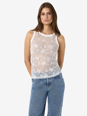 Kvinde iført hvid ærmeløs top med blonder og blomster og blå jeans, stående med hænderne bag ryggen mod en ensfarvet baggrund.