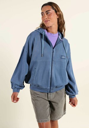 Femme portant un sweat à capuche bleu à fermeture éclair sur un t-shirt lavande et un short gris à hauteur des genoux, debout devant un fond clair uni.