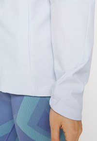 Gros plan sur la main d'une personne portant une veste blanche à manches longues et des leggings bleus à motifs, montrant la texture du tissu et les détails de la couture.