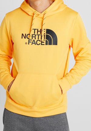 Osoba nosząca żółtą bluzę z kapturem z czarnym logo "The North Face" i sznurkami, ręce w przedniej kieszoni, w połączeniu z szarymi spodniami.