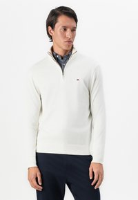 Mann som har på seg en hvit quarter-zip genser over en mønstret skjorte med knapper og mørke bukser, stående mot en ensfarget hvit bakgrunn.