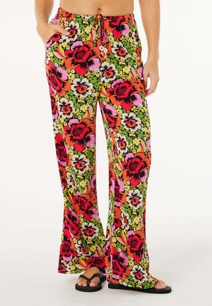 Femme portant un pantalon ample à imprimé floral vif en rouge, rose, jaune et vert, associé à des tongs noires.