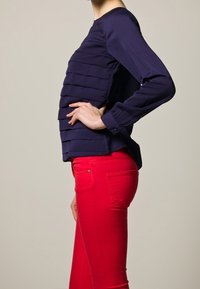 Blusa azul marino de manga larga con pliegues horizontales texturizados, combinada con jeans rojo ajustados. El conjunto presenta un acabado liso y un diseño entallado.
