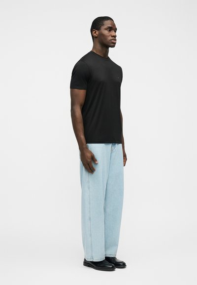 Άντρας στέκεται στραμμένος προς τα δεξιά φορώντας μαύρο πουκάμισο με κοντά μανίκια, ανοιχτό μπλε τζιν με φαρδιά πόδια και μαύρα παπούτσια στο λευκό φόντο.