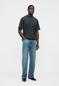 Man staat met één hand in zijn zak, draagt een donkergrijs T-shirt met korte mouwen, lichtblauwe jeans met rechte pijpen en zwarte puntige schoenen.