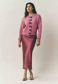 Rosa ulltröja med svarta slingsknappar, tillsammans med en lång, glänsande burgundysatin kjol. Modell som bär svarta sandaler med remmar.