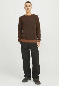 Pulover tricotat maro cu un model texturat, manșete cu nervuri și pantaloni cargo negri cu buzunare laterale și o croială lejeră, asortați cu pantofi închiși la culoare.