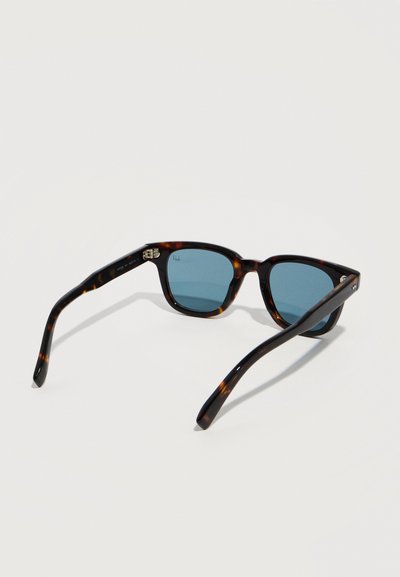 Γυαλιά ηλίου με καφέ ταρταρούγα πλαίσιο και ορθογώνια μπλε φακούς, ανοιχτά και τοποθετημένα σε λευκή επιφάνεια.